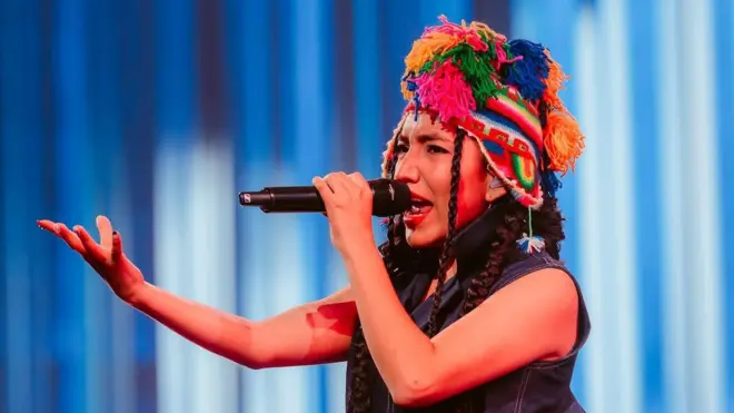 Renata Flores con un chullo de colores, gorro típico de Perú, con un micrófono en la mano y cantando.