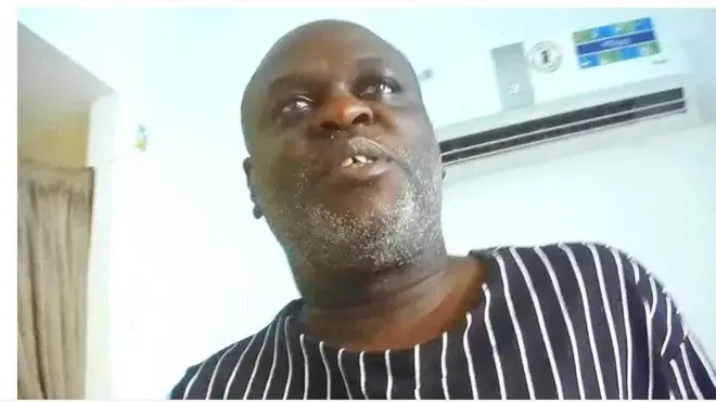 Aworan yii ṣafihan Charles Mwegiswa, eyin rẹ meji han nita, o n wo ọọkan, o wọṣo dudu to ni ila funfun tẹẹrẹtẹ