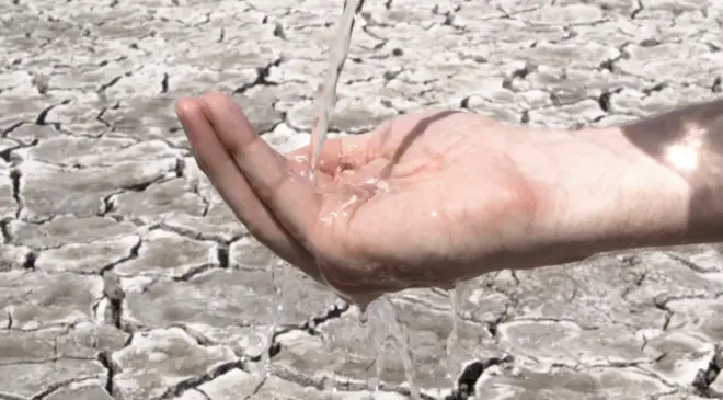 පොළව ඉරිතලාගිය පසුබිමක පුද්ගලයකුගේ අතට ජලය වැටෙන අයුරු දැක්වේ.