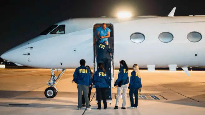 Mavi formalı bir adam elleri kelepçeli bir jetten iniyor, 4 FBI memuru arkaları dönük şekilde yerde onu karşılıyor. 