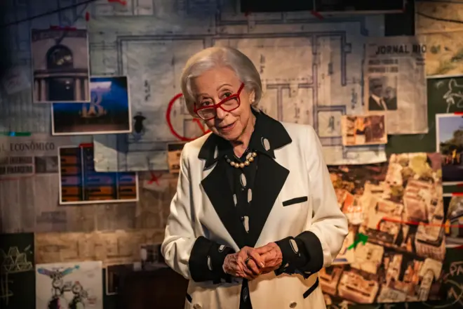 Fernanda Montenegro, de cabelos brancos e usando óculos de armação vermelha, aparece em primeiro plano diante de um painel cheio de fotos, recortes de jornal e um mapa com marcações em vermelho, como em uma parede de investigação. Ela veste um blazer branco com detalhes pretos, camisa escura com estampa clara e um colar de pérolas, mantendo as mãos juntas à frente do corpo e olhando para a câmera com expressão inquisitiva.