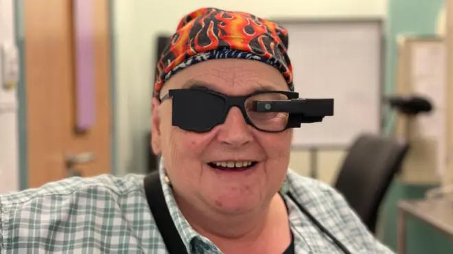 希拉·歐文直視鏡頭——她戴著一副黑色眼鏡，左眼正前方的一個鏡片上裝著一個攝影機。右鏡片則完全是黑色的。她穿著格子襯衫，戴著一條色彩鮮豔的頭巾。