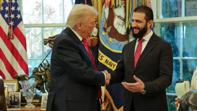ホワイトハウスの大統領執務室の窓際でにこやかに握手するトランプ氏とアル・シャラア氏。共にダークスーツに赤いネクタイを着けている。アル・シャラア氏は長いひげを蓄えている