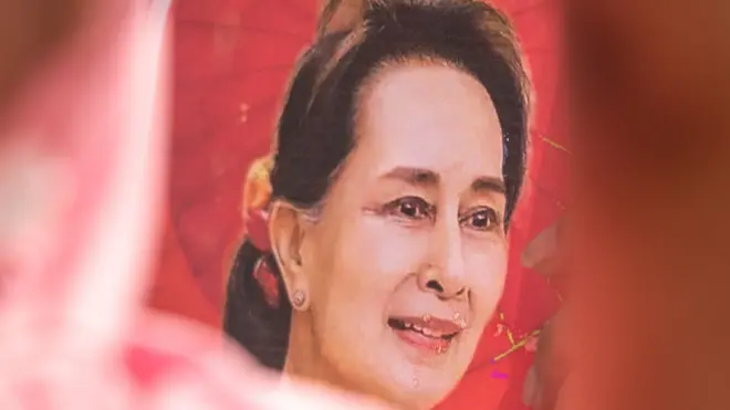 A photo of Aung San Suu Kyi on a red banner held up by crowds demonstrating in the Thai capital, Bangkok, on 1 February 2023. The left cheek of a woman wearing a red and white headscarf is out of focus in the foreground to the left of the banner. 
