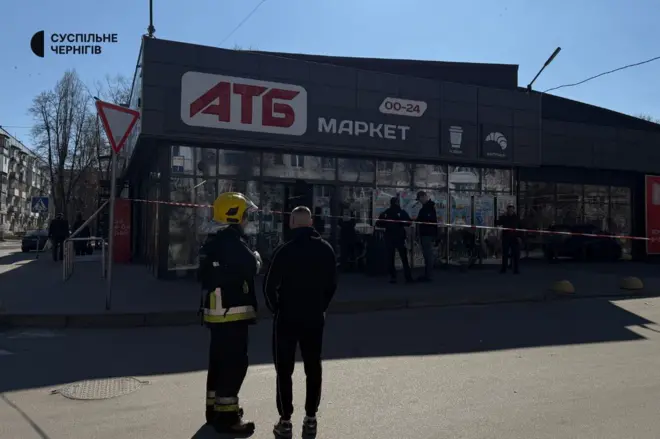 Будівля супермаркету «АТБ», перед входом до якого натягнута червоно-біла стрічка, що обмежує територію. Біля стрічки стоять кілька людей, серед них — рятувальник у захисному спорядженні. На зображенні також є логотип «Суспільне Чернігів».