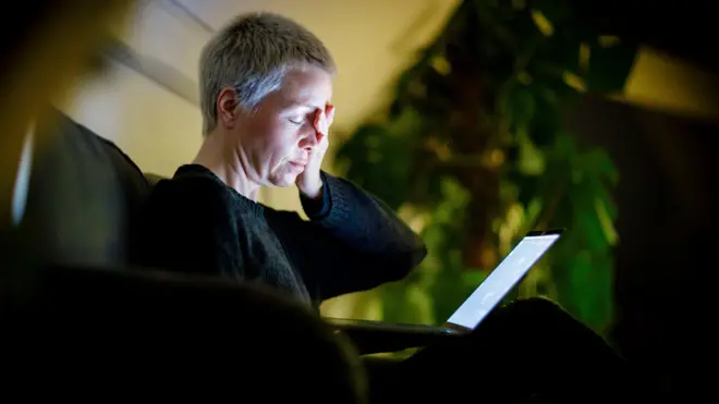 Уставшая женщина с короткой стрижкой с закрытыми глазами сидит на диване с ноутбуком на коленях. Левую руку она приложила к лицу.