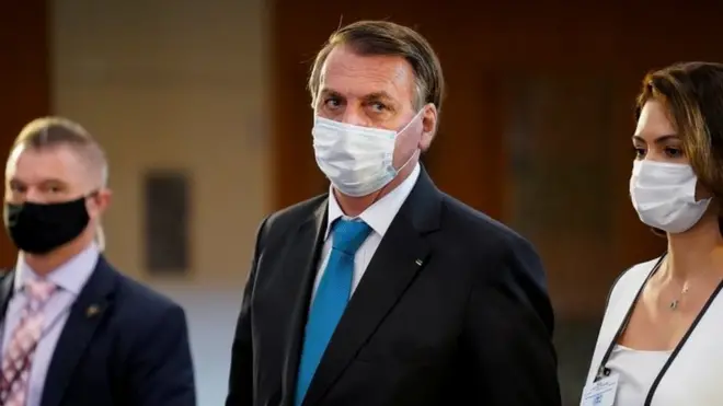 Jair Bolsonaro ao lado da primeira-dama Michele, ambosbetano aposta de futebolmáscara e sendo observados por um segurança ao fundo