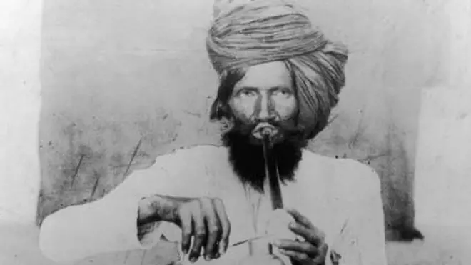 A man in a turban sits cross-legged smoking opium in Meywar, Rajputana
