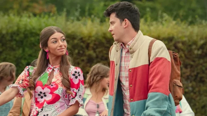 Mimi Keene as Ruby and Asa Butterfield as Otis in Sex Education. Ruby and Otis are speaking to each other outside as they wheel their bikes. Ruby, a young woman with long light brown hair, wears a short floral dress and large pink earrings. Otis, a young man with short dark hair, wears orange cord trousers, a pink plaid shirt and a blue, cream and red striped puffer jacket.