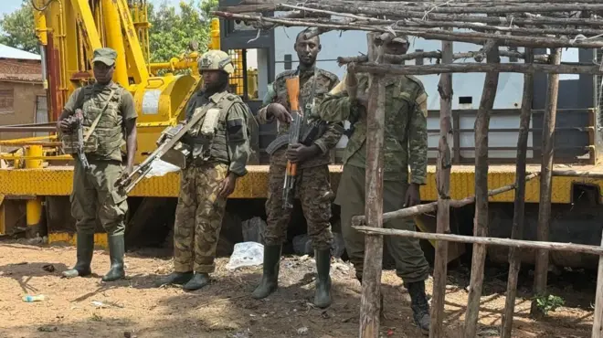 Abarwanyi bane ba M23 bahagaze imbere y'ikimodoka, umwe muri bo ahagaze musi y'inzu y'ibiti itari bwaheze kwubakwa