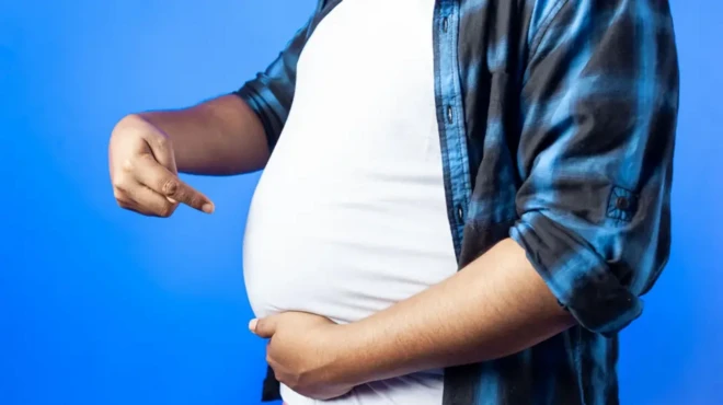 رجل يرتدي قميص داخلي أبيض وفوقه قميص أسود وأزرق يشير بإصبعه إلى كرشه المنتفخ