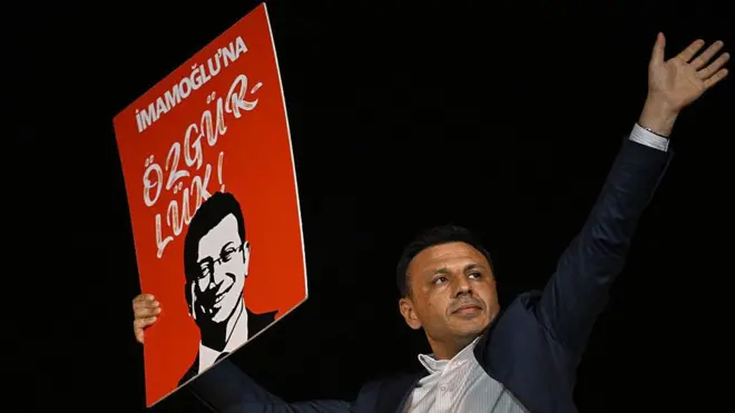 Özgür Çelik elinde "İmamoğlu'na Özgürlük" yazılı bir döviz tutuyor ve sol kolunu yukarıya kaldırıyor