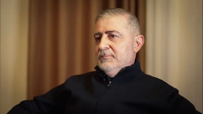 يجلس رجل على كرسي مرتدياً سترة سوداء وينظر إلى جانب الكاميرا