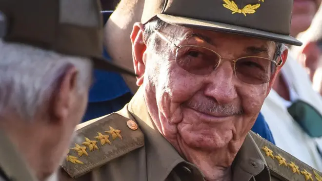 Primer plano de Raúl Castro, con gafas de sol y uniforme, sonriendo a otra persona