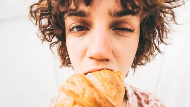一名女子咬下一口牛角面包,同时眨了眨一只眼睛。鱼眼镜头拍摄的镜头夸张地突出了她的面部表情、牛角面包以及咬合的动作。