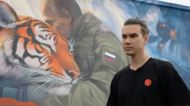 Filipp Dulmachenko stands in front of his mural of Vladimir Putin hugging a Siberian tiger