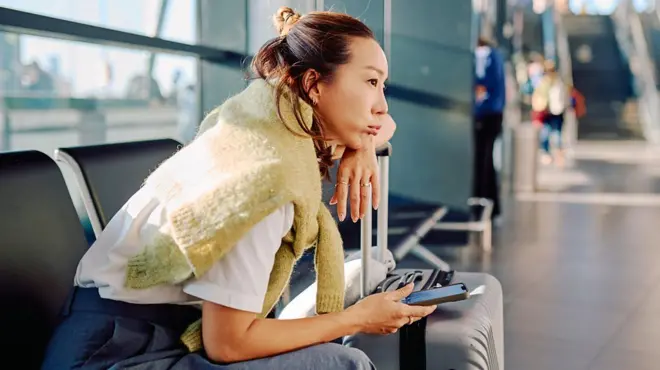 مسافرة تنتظر في المطار، تتكئ على حقيبتها وتحمل هاتفاً في يدها.