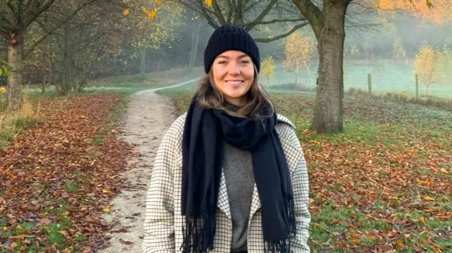A woman with brown hair in a black beanie and black scarf stands in an autumnal landscape with fog, trees and brown leaves