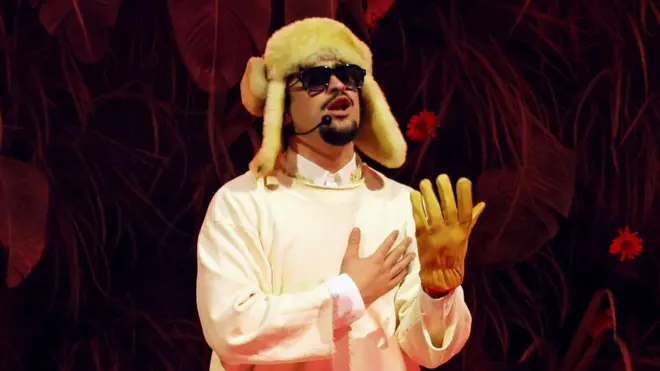 Bad Bunny cantando sobre un escenario durante su residencia en Puerto Rico luciendo un gorro esquimal, gafas oscuras y un solo guante.