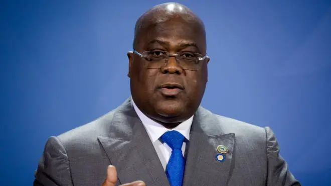Perezida wa DR Congo Félix Tshisekedi yafotowe ubwo yari ari mu kiganiro n'abanyamakuru i Berlin mu Budage, ku itariki ya 27 Kanama (8) mu 2021, mu nama yahuje itsinda ry'ibihugu bikize cyane rya G20 n'Afurika. Yambaye indorerwamo, ikositimu iri hagati y'amabara ya khaki n'ikigina, ishati y'umweru na karuvati y'ubururu, akoze ikimenyetso n'ikiganza. 