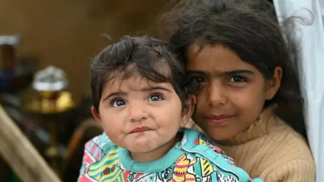 ایک بے گھر لڑکی جس نے بیج رنگ کا جمپر پہن رکھا ہے، اپنی چھوٹی بہن کو اٹھائے ہوئے ہے۔ دونوں کی بڑی بھوری آنکھیں اور الجھے ہوئے بال ہیں۔ وہ لبنان کے دارالحکومت بیروت میں بے گھر افراد کے ایک غیر رسمی کیمپ میں ایک خیمے کے داخلی دروازے پر کھڑی ہیں۔