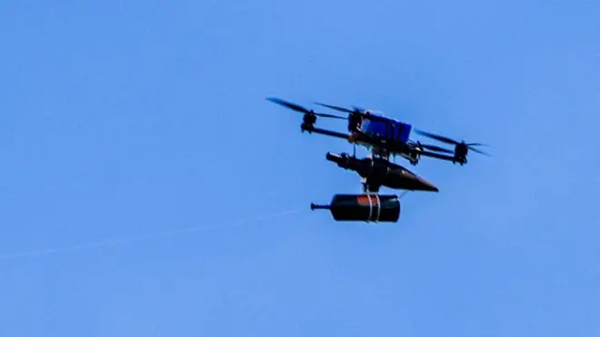A fibre-optic drone flies in a clear blue sky across the border from Lebanon into Israel. It has a spool of fibre optic wire handing under a chassis, supported by four propeller blades around a blue rectangular battery.