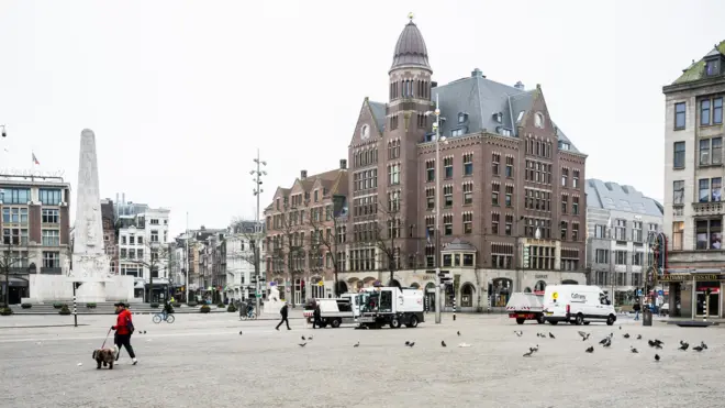 На площади Дам в Амстердаме видны обелиск, голуби, человек с собакой на поводке, белый фургон и уборочная машина