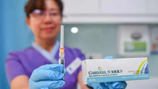 A healthcare worker in purple uniform and blue medical gloves holds a syringe toward the camera while holding a white vaccine box in the other hand
