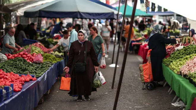 Pazarda yürüyen bir kadın, ellerinde poşetler taşıyor. Sağ ve sol tarafındaki tezgahlarda meyve ve sebzelere bakan başka kadınlar görülüyor