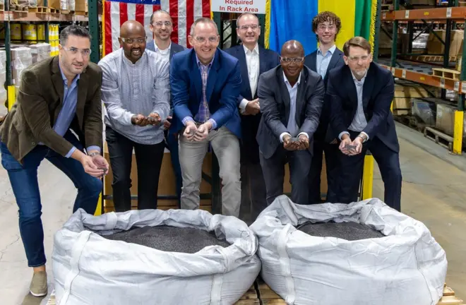 Ambasaderi w'Amerika i Kigali (hagati) akikijwe n'abandi bagabo bafite amabuye y'agaciro mato y'umukara mu biganza, bayafatiye hejuru y'imifuka ibiri y'umweru irimo andi mabuye y'agaciro. Barareba muri 'camera', inyuma yabo hari ibendera ry'u Rwanda n'iry'Amerika. avuga ko ari umunsi w'amateka kuba u Rwanda rwohereje bwa mbere aya mabuye y'agaciro muri Amerika