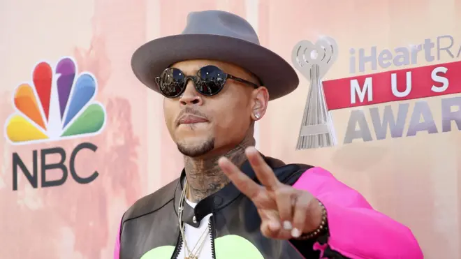 Chris Brown pose on di red carpet for di IHeartRadio Music Awards. E wear one black jacket wit pink sleeves and e dey do di peace sign wit im left hand.E wear one grey hat and sunglasses.