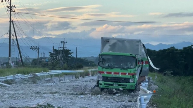 地震で道路の舗装がはがれガードレールが曲がっている。停めてあるトラックも荷台が大きく損傷している