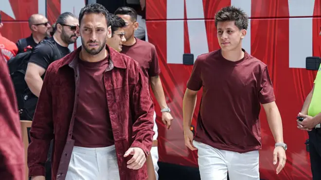 Arda Güler ve Hakan Çalhanoğlu yürüyor. İkisi de aynı kıyafeti giymiş, bordo yoğun bir rahat takım vu. Arkada takım otobüsü var.