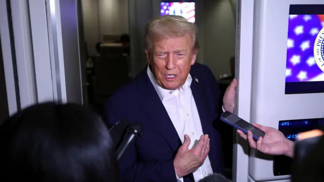 Donald Trump, de terno azul marinho e camisa branca, parado numa porta, conversa com repórteres que apontam microfones e celulares