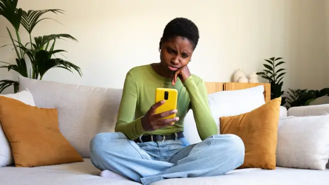 One young woman sit down ontop chair and dey look her smartphone wit tight face as she dey vex.