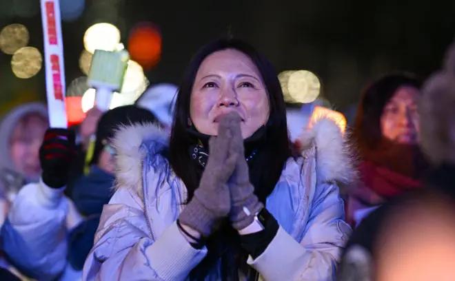 A woman wearing grey gloves stands with her hands clasped together against a night time backdrop