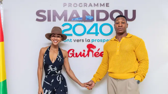 Meagan Good (à droite) vêtue d'une combinaison noir et blanc et d'un fedora marron, et Jonathan Majors (à gauche) vêtu d'un pull jaune et d'un pantalon beige se tiennent la main devant une pancarte sur laquelle on peut lire « Programme Simandou 2040 ». Un drapeau guinéen est déployé à l'extrême droite du cadre