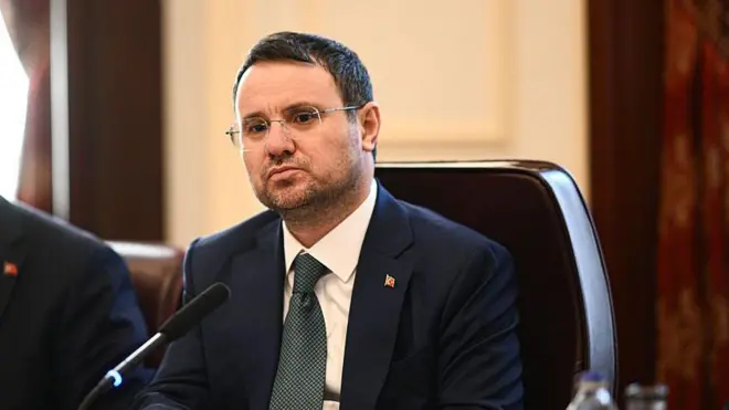 Adalet Bakanı Akın Gürlek oturuyor, önünde mikrofonlar var. 