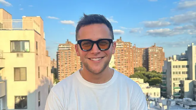 Retrato colorido de Andrew Greene, um homem branco jovem, usando uma camiseta branca, óculos de lente amarela e com uma cidade ao fundo