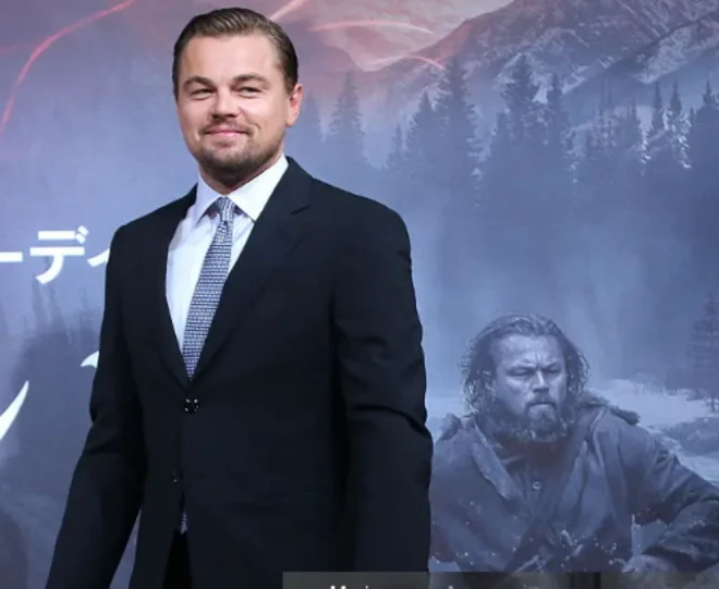 Leonardo DiCaprio attends the press conference for The Revenant film at the Ritz Carlton on 23 March 2016 in Tokyo, Japan. He is wearing a dark suit, white shirt with a blue tie. Behind him is a poster promoting the film, showing DiCaprio in character as High Glass. Glass is depicted as a man with unkempt hair, wearing a heavy tunic. 