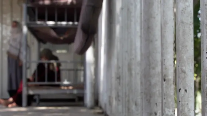 Metal bars as part of a gate to a facility. A woman in the distance sits on a metal bunk bed. Another woman stands next to her, with her face and head covered.