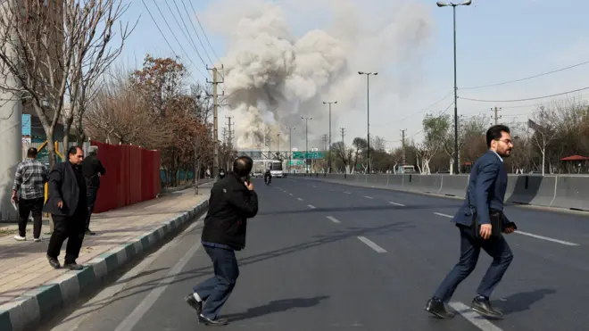 Міська вулиця, по якій люди швидко відбігають убік, озираючись назад. На горизонті видно велику хмару густого диму, що піднімається над дорогою — ймовірно, від вибуху чи удару. Узбіччям рухаються перехожі, а проїжджа частина майже порожня.