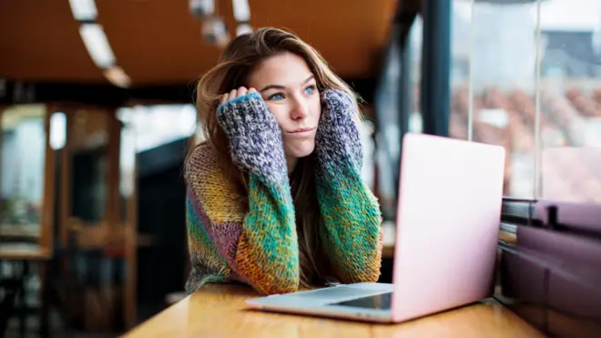 Una joven aburrida mira por la ventana. Está sentada en una biblioteca, usando una computadora portátil.