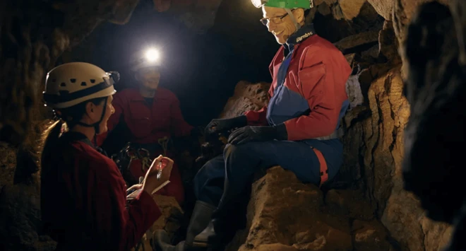 Três pessoas dentrocupom casa de apostauma caverna, usando capacetes com lanternas. Um homemcupom casa de apostamacacão azul está sentado, conversando com os outros dois, que estãocupom casa de apostapé segurando objetos.