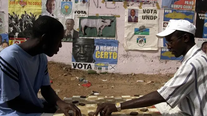 Des résidents de Bissau s'adonnent au jeu de dame devant un mur couvert d'affiches de partis politiques (illustration)