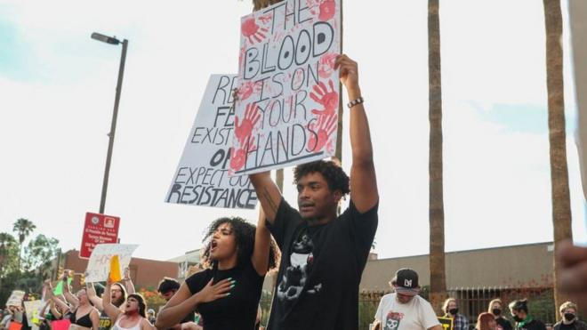 Fotografia colorida mostra um homem e uma mulher negros protestando com cartazes e diversas pessoas brancas protestandopoker online brasilsegundo plano