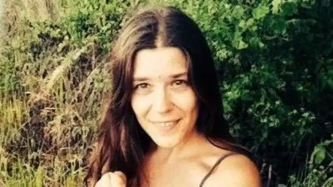 Nina olha para a câmera e sorri. Atrás dela há várias árvores