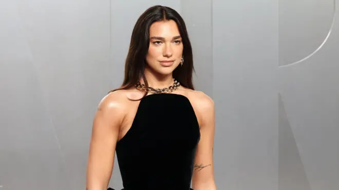 Dua Lipa pewnie pozuje przed kamerą, ubrana w czarną sukienkę z gorsetem i złoty, gruby naszyjnik. Jej długie, ciemne włosy są rozpuszczone.