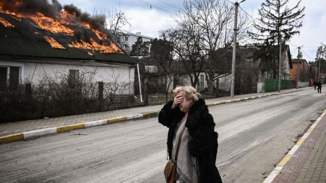 A woman covers her face with her hand as she stands in front of a house burning in Irpi, outside Kyiv, 4 March 2022