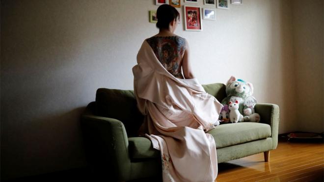 A woman kneels on a couch with a garment pulled down to reveal her large back tattoo to the camera